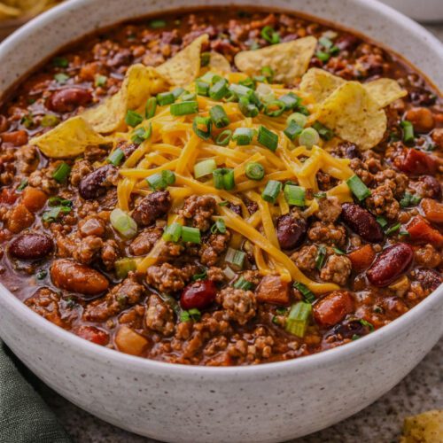 Hearty bowl of homemade chili topped with cheddar cheese, scallions, and corn chips