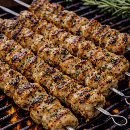 Grilled yogurt-marinated chicken skewers with char marks cooking over open flame with lemon and herbs.