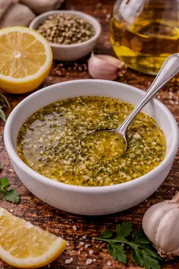 Greek ladolemono sauce in a small bowl with olive oil, lemon juice, garlic, and oregano, surrounded by fresh lemons and herbs on a wooden surface.