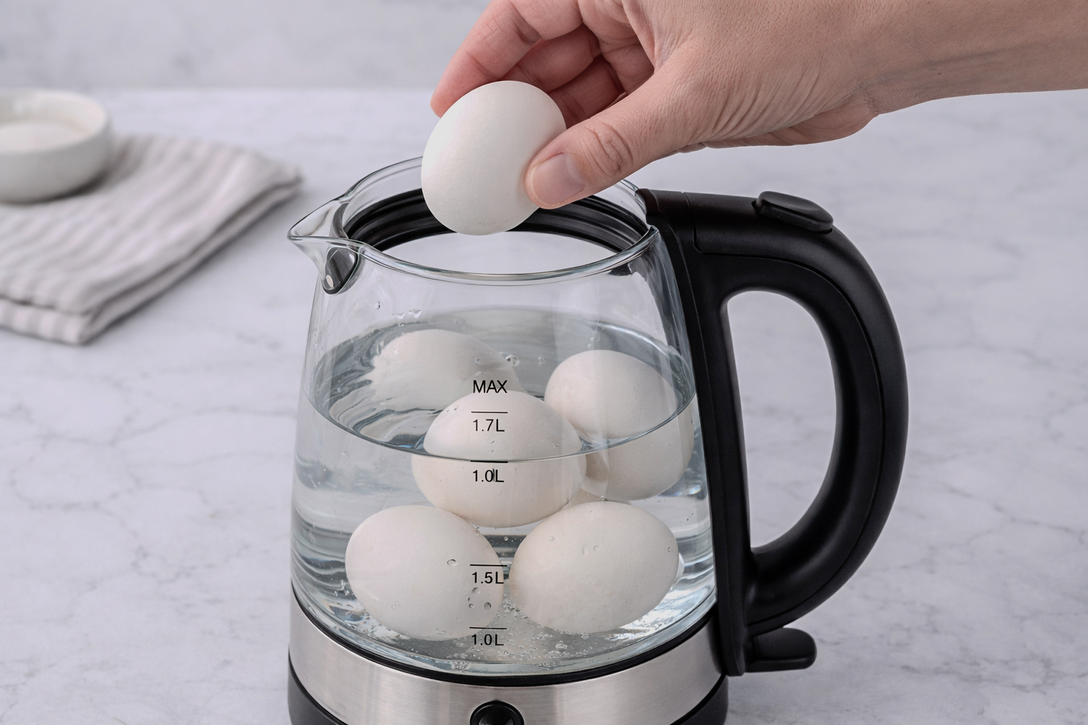 Hand placing raw eggs into a glass electric kettle filled with water on a kitchen counter.