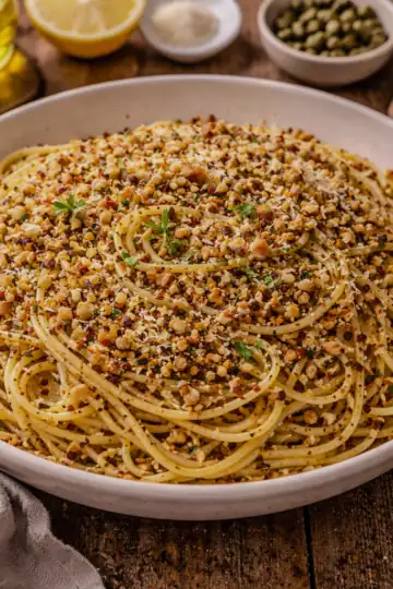 Plate of walnut breadcrumb pasta served on a rustic wooden table with garlic, olive oil, herbs, and bread in the background.