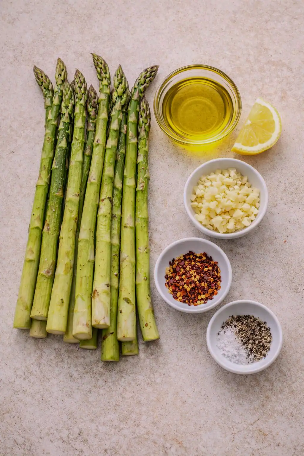 Fresh asparagus spears arranged on a countertop with olive oil, lemon, minced garlic, red pepper flakes, salt, and black pepper in small bowls, ready for grilling