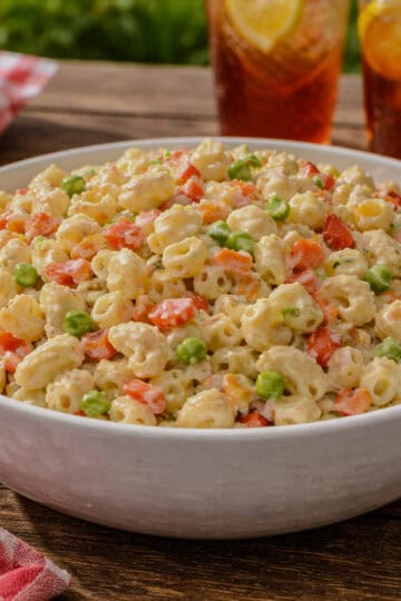 A bowl of creamy macaroni salad with shell pasta, diced bell peppers, peas, and carrots sitting on a wooden picnic table with a red checkered cloth and summer picnic foods in the background.