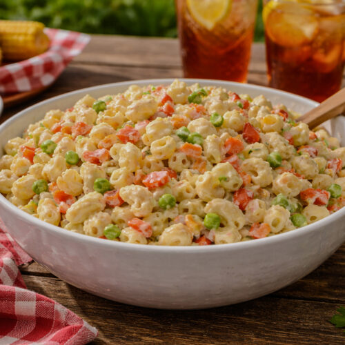 A bowl of creamy macaroni salad with shell pasta, diced bell peppers, peas, and carrots sitting on a wooden picnic table with a red checkered cloth and summer picnic foods in the background.