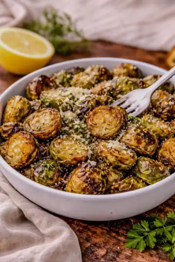 Crispy roasted Brussels sprouts in a white bowl topped with grated cheese and herbs, served with a lemon wedge on a wooden table.