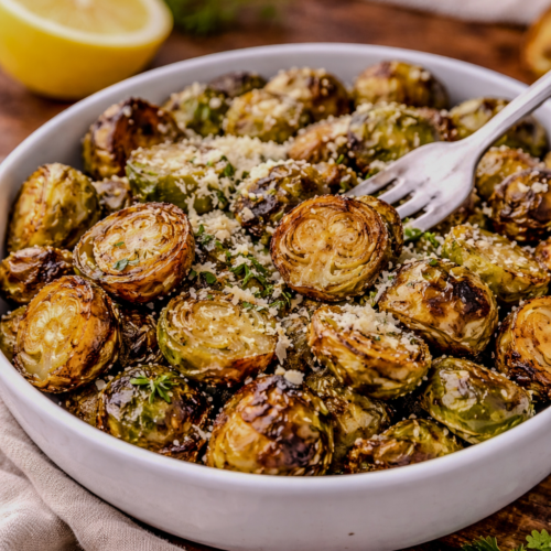 Crispy roasted Brussels sprouts in a white bowl topped with grated cheese and herbs, served with a lemon wedge on a wooden table.