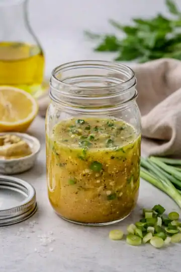 Rice vinegar Dijon dressing in a glass mason jar on a kitchen counter with chopped scallions, olive oil, and lemon nearby.