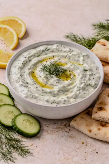 Creamy tzatziki in a white bowl garnished with olive oil and fresh dill, surrounded by cucumber slices, lemon wedges, and warm pita on a light countertop