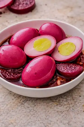 Pickled red beet eggs in a shallow bowl with sliced beets and pink brine, including halved eggs showing bright yellow yolks