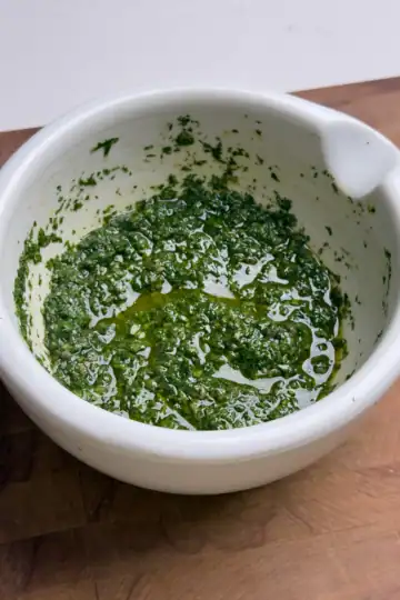 A vibrant green zhoug sauce in a rustic bowl, made with fresh cilantro, garlic, and olive oil, with a slightly textured consistency and herbs scattered on a wooden surface.