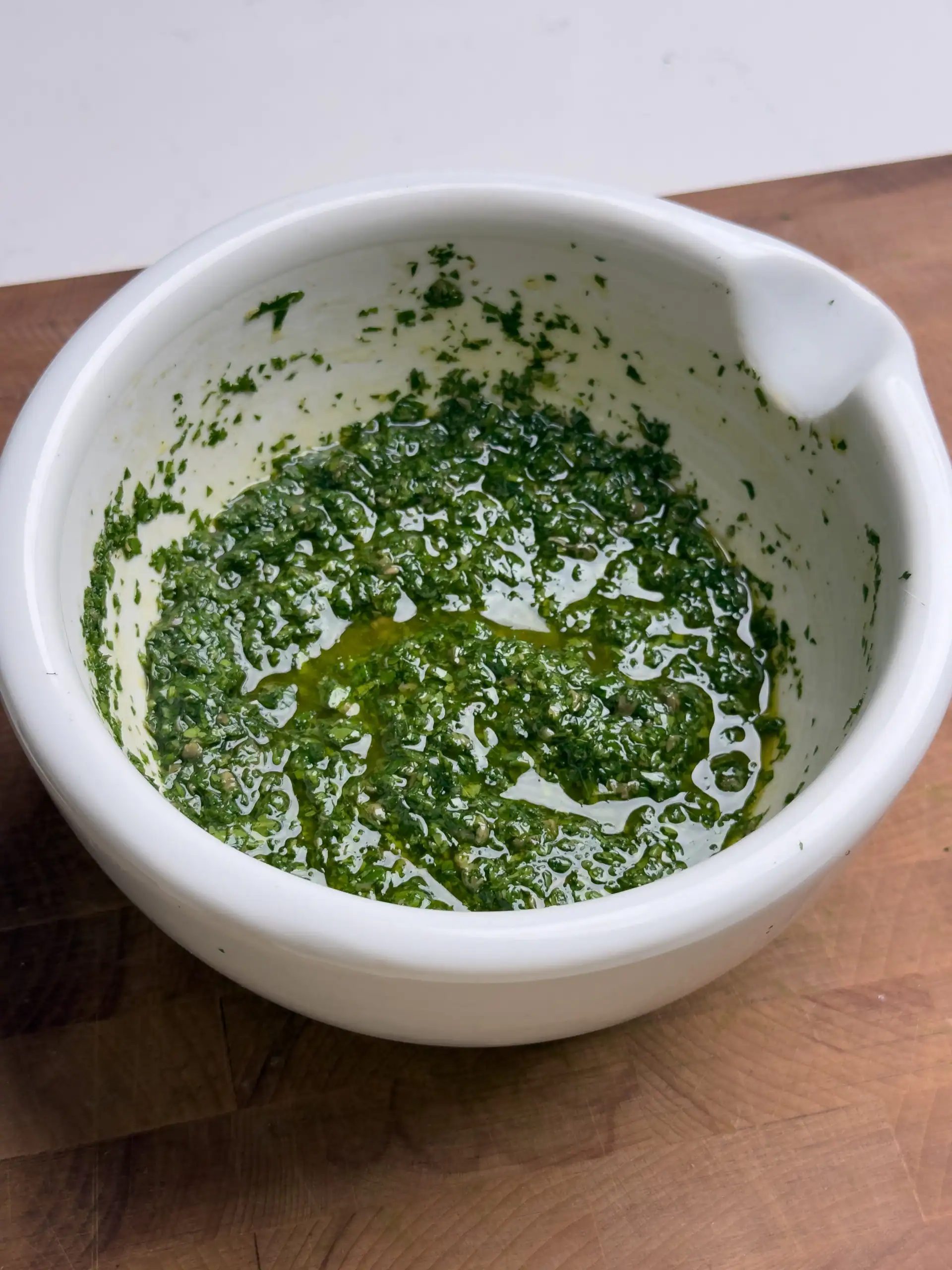 A vibrant green zhoug sauce in a rustic bowl, made with fresh cilantro, garlic, and olive oil, with a slightly textured consistency and herbs scattered on a wooden surface.