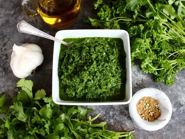 All the ingredients for zhoug sauce arranged on a counter, including fresh cilantro, garlic cloves, jalapeño, whole spices, and olive oil.