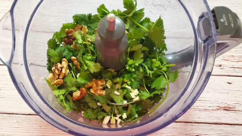 Fresh cilantro, garlic, jalapeño, and spices added to a food processor bowl before blending.