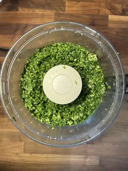 Olive oil being drizzled into a food processor as a green herb sauce blends into a smooth, slightly textured consistency.